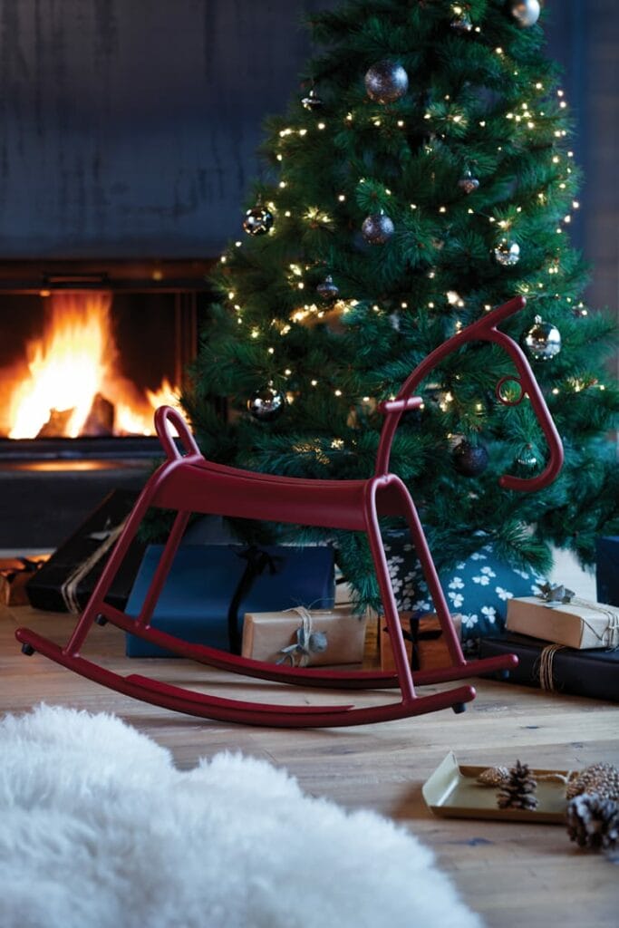 Un Cheval à Bascule Adada rouge devant un sapin de Noël décoré d'ornements bleus et argentés, avec une cheminée allumée en arrière-plan.