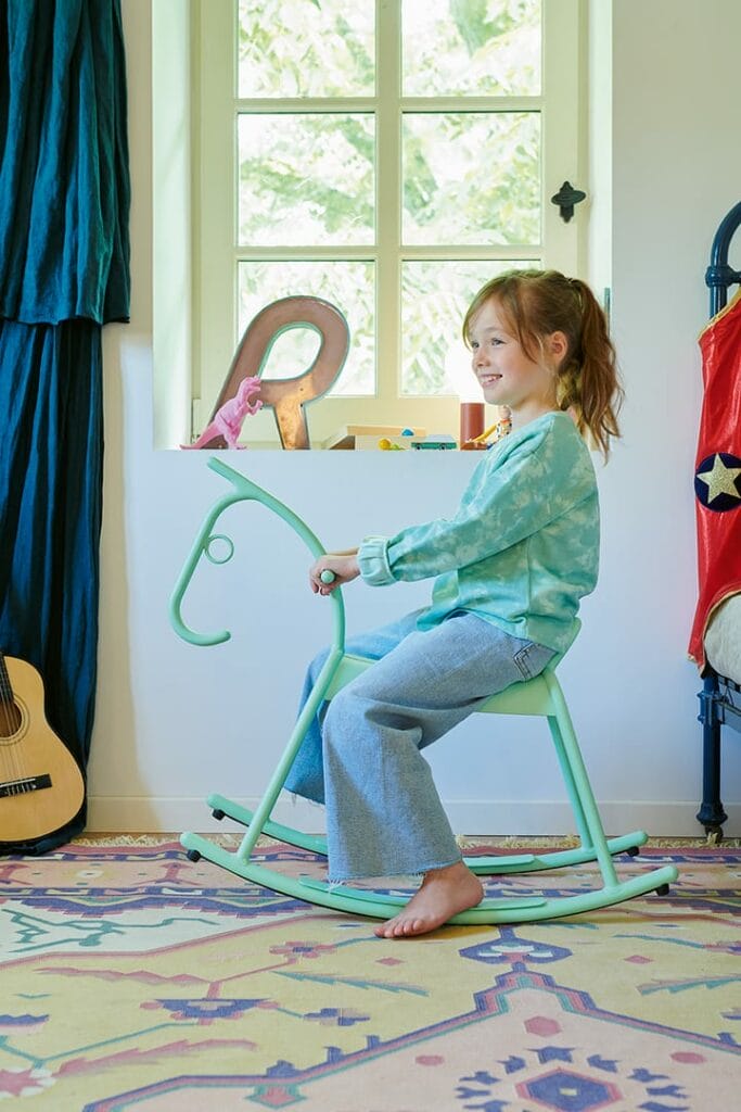 Une jeune fille vêtue de vêtements décontractés sourit assise sur un Cheval à Bascule Adada vert dans une pièce colorée avec une fenêtre, des rideaux et des jouets.