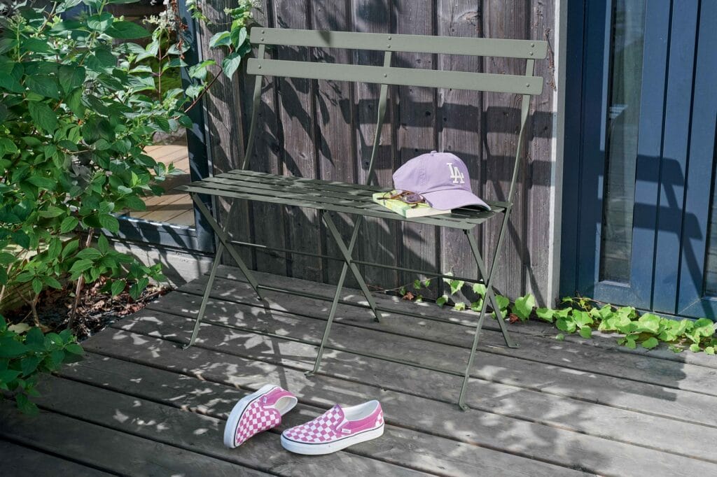 Une Banquette Bistro 2 places en métal vert avec une casquette et un livre ouvert à côté de chaussures à carreaux roses et blancs, située sur une terrasse en bois au milieu d'une verdure dense et d'une porte bleue.