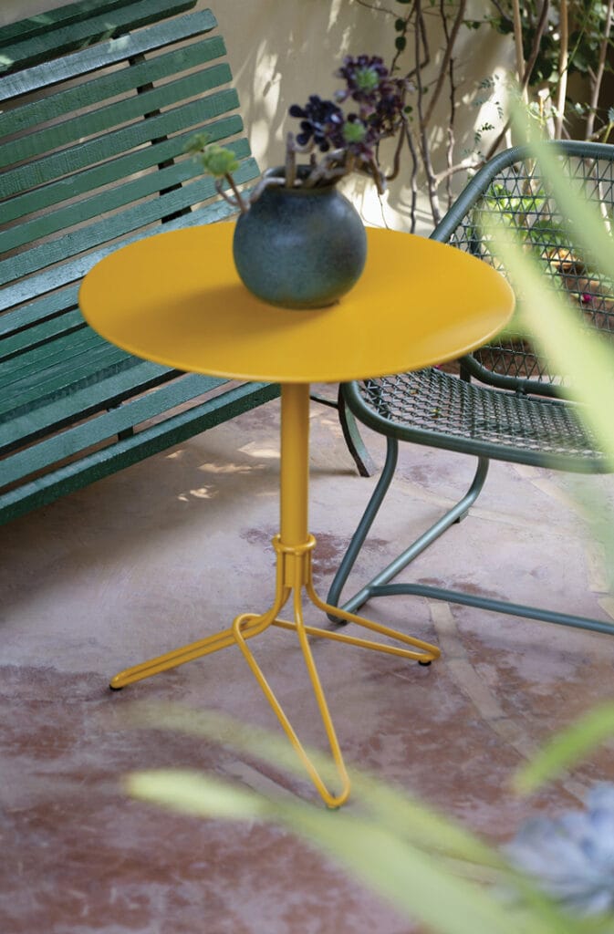 Une Fleur Guéridon ronde en métal jaune avec un vase bleu et de la verdure sur un patio en terre cuite, entouré de plantes et d'une chaise verte.