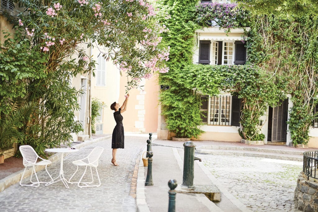 Une femme prend une photo de vignes en fleurs surplombant une charmante rue pavée, avec des chaises blanches et une fleur de Guéridon ronde à proximité.
