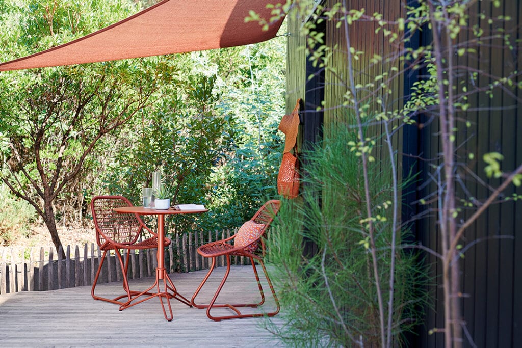 Patio extérieur avec deux chaises orange et une table Guéridon Flower sous une voile d'ombrage, entouré de verdure et d'une clôture noire moderne.