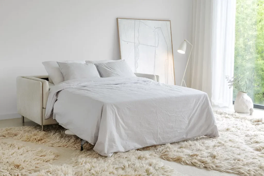 Chambre moderne avec un lit défait, un tapis moelleux, un lampadaire et des œuvres d'art abstraites près d'une fenêtre avec des rideaux blancs et un Canapé Pixie supplémentaire.