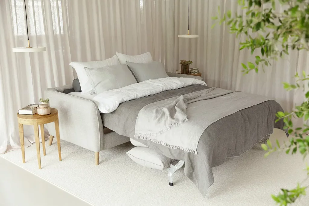 Une chambre moderne et confortable dotée d'un lit soigneusement fait avec une literie grise et blanche, flanqué de tables de chevet en bois et de rideaux moelleux, rehaussé par l'ajout élégant d'un Canapé Lucy.