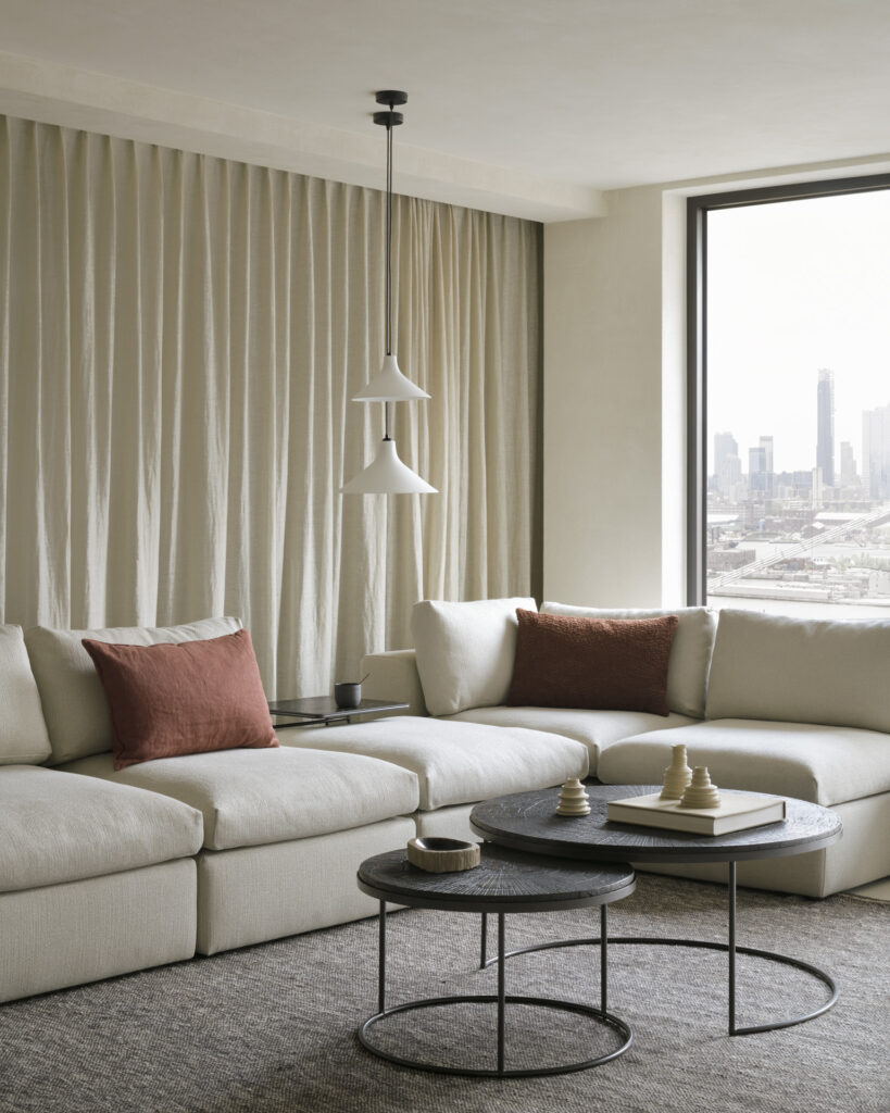 Un salon moderne avec un canapé sectionnel beige clair, deux coussins Slice Umber couleur rouille et une table basse Nesting - Minerals - Slice Umber - ronde - ensemble de. Une suspension est suspendue au-dessus, tandis qu'une grande fenêtre révèle le paysage urbain à l'extérieur. Des rideaux neutres recouvrent le mur.