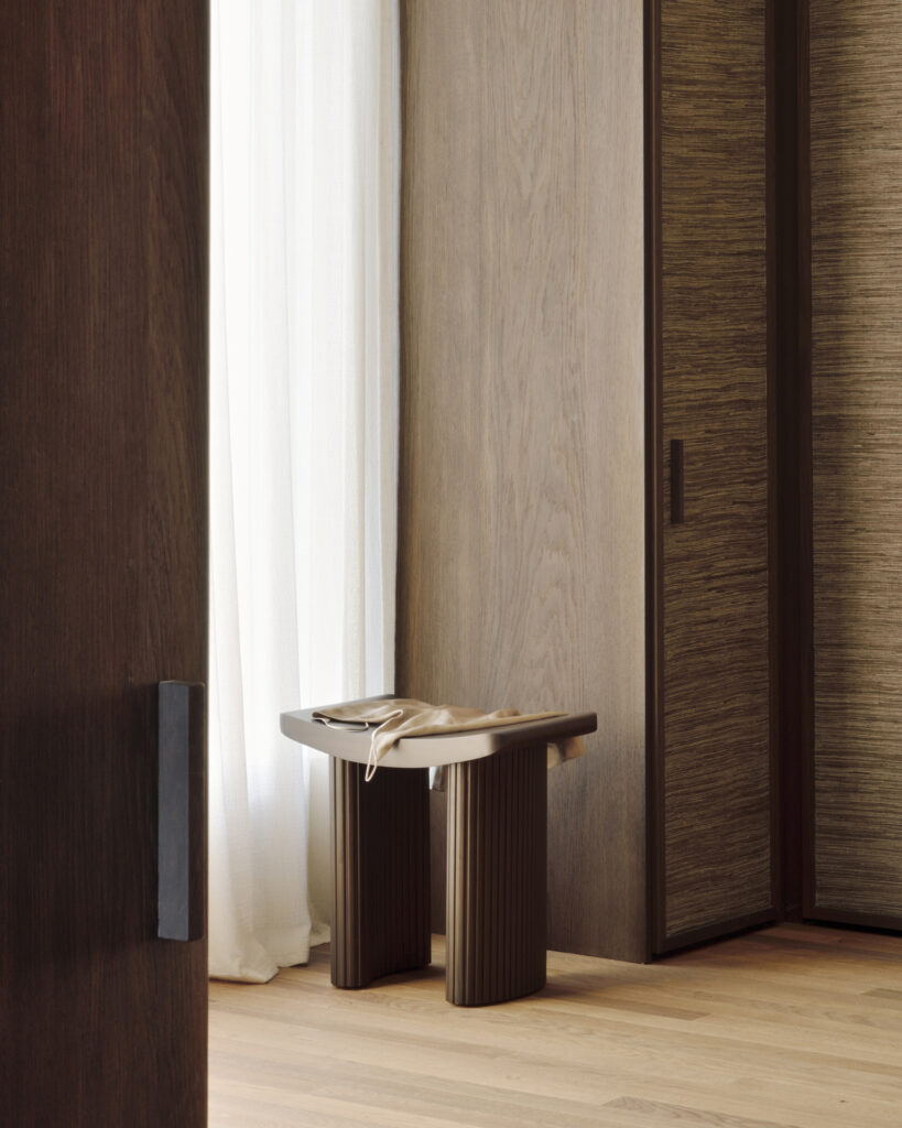 Un tabouret minimaliste Roller Max en bois - acajou verni - marron foncé avec tissu plié est placé devant un rideau blanc et des lambris en bois, posés sur un parquet en bois clair.