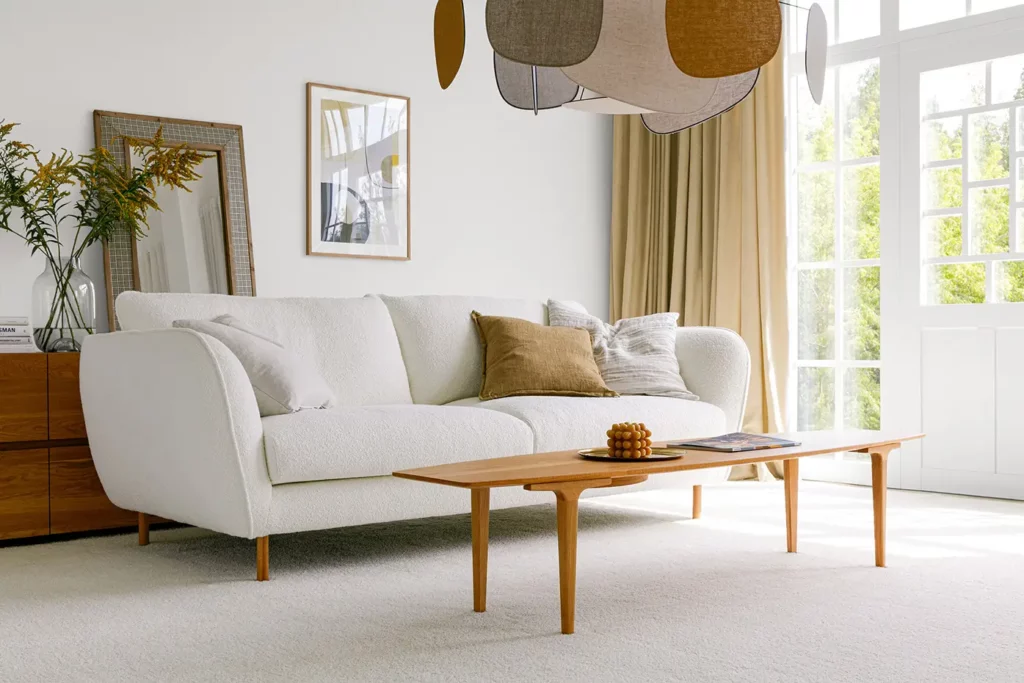 Un séjour lumineux avec un Canapé Emma 3 places Lux LCV - Willow 1 blanc cassé, une table basse en bois, de grandes fenêtres et une plante en pot.