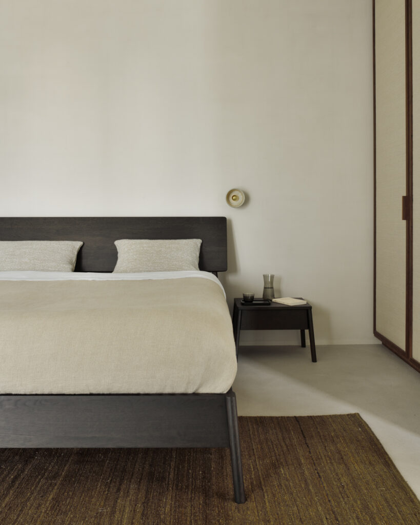 Dans une chambre minimaliste, un lit Air en chêne verni foncé sert de point central. Cette pièce maîtresse élégante est dotée d'un matelas 160x200 qui allie confort et élégance. La chambre dispose d'une literie de couleur claire, complétée par une petite table d'appoint et un tapis tissé sur le sol aux tons clairs. Les murs et les armoires de couleur neutre contribuent à l'ambiance sereine, tandis qu'une petite applique murale fournit un éclairage doux à proximité.