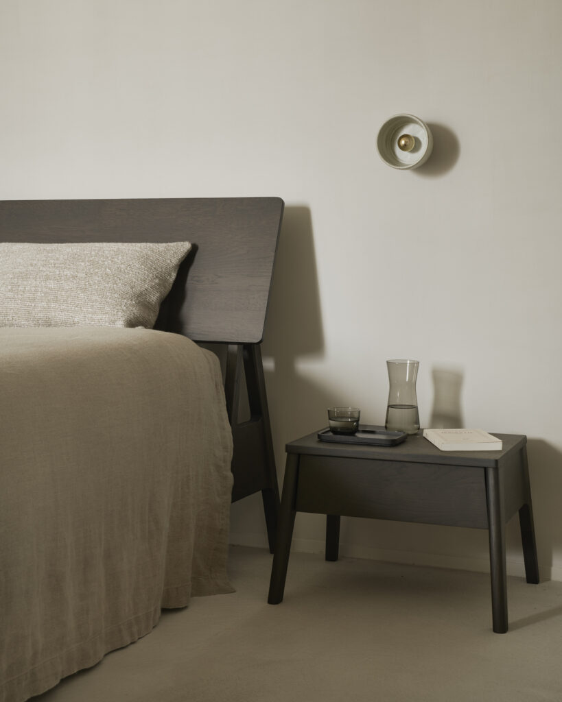 Une chambre minimaliste présente un lit en bois foncé avec une literie beige. La table de chevet Air en chêne verni, dotée d'un tiroir, supporte une carafe, un verre et un livre. Une applique murale illumine le mur neutre.