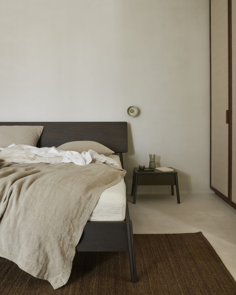 Une chambre minimaliste présente un lit en bois avec une literie beige, complété par la table de chevet Air en chêne verni, avec une teinte marron et un petit tiroir pouvant contenir un livre et des lunettes. Un éclairage doux est fourni par un luminaire mural au-dessus.