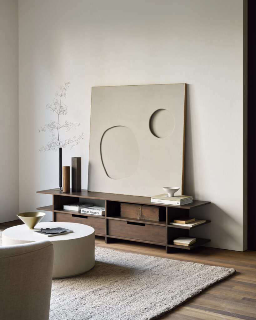 Salon minimaliste avec une grande œuvre d'art abstrait sur un Meuble TV Kabuki. Une table basse ronde, un tapis beige et des objets de décoration dont des livres et des vases complètent l'espace.