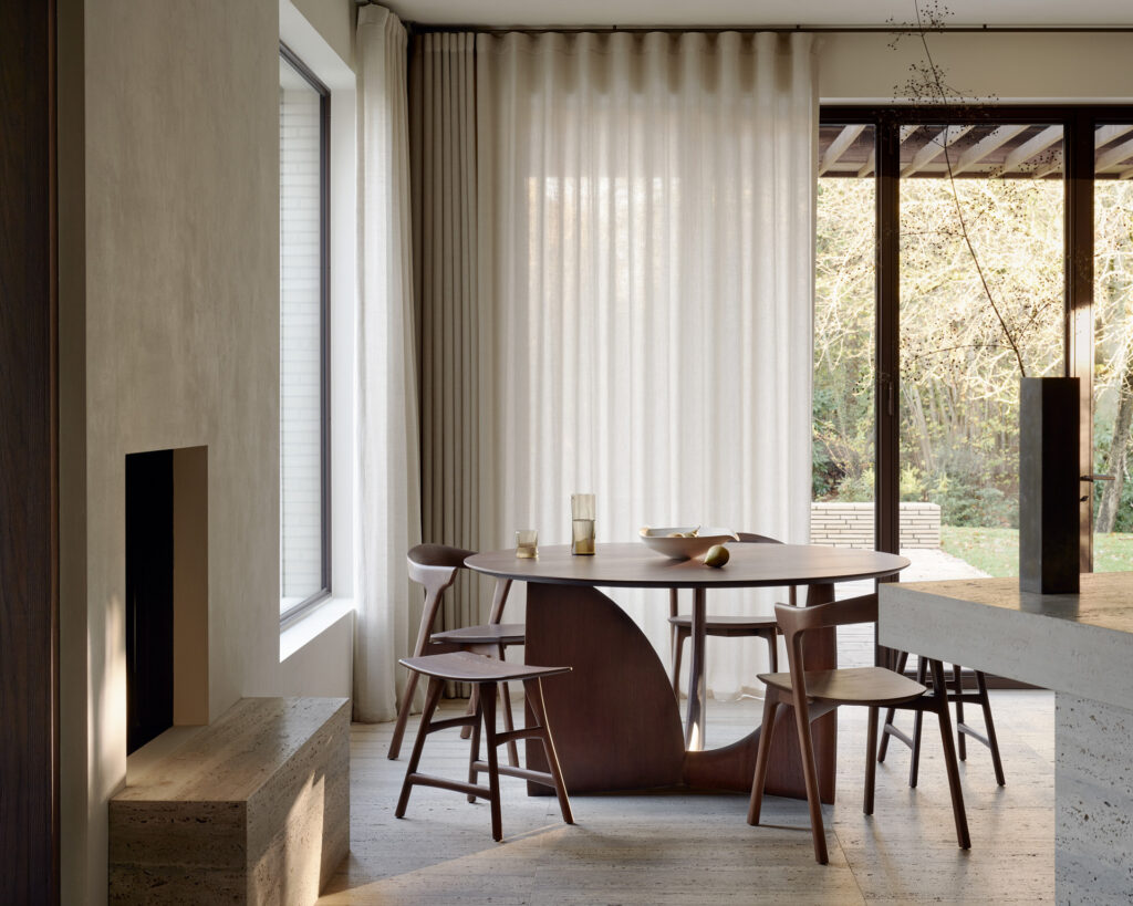 Une salle à manger moderne avec une table à manger géométrique - teck vernis - marron - ronde, cinq chaises assorties, de grandes fenêtres avec voilages et un vase avec des branches sur la table. La pièce présente un décor minimaliste et une lumière naturelle, soulignant la simplicité élégante de l'espace.