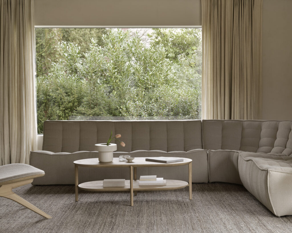 Un salon moderne comprend un canapé sectionnel 3 places Écru Eco-friendly Canapé N701, une table basse ronde ornée d'une plante et d'un livre, une chaise longue et une grande fenêtre avec des rideaux beiges donnant sur la verdure.