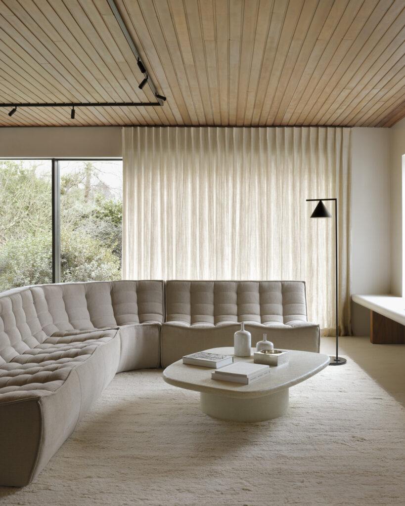 Un salon moderne comprend un canapé sectionnel beige avec un module d'angle arrondi, une table basse ovale écologique écrue avec des objets décoratifs, un lampadaire, de grandes fenêtres avec des rideaux lumineux et un plafond en bois.