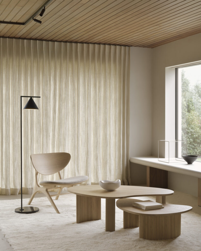 Un salon minimaliste avec une chaise en bois, des tables basses rondes en chêne naturel, un lampadaire et une décoration aux couleurs neutres sur fond de rideaux beiges et d'une grande fenêtre.