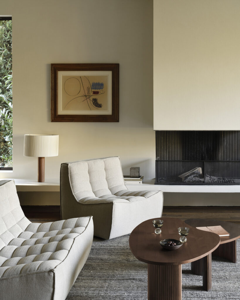 Un salon moderne avec des chaises rembourrées blanches, une table basse (table basse) en teck teinté marron, une cheminée et une peinture abstraite encadrée au mur. Une petite lampe de table est placée près de la fenêtre, sa lumière créant une lueur chaleureuse et invitante.