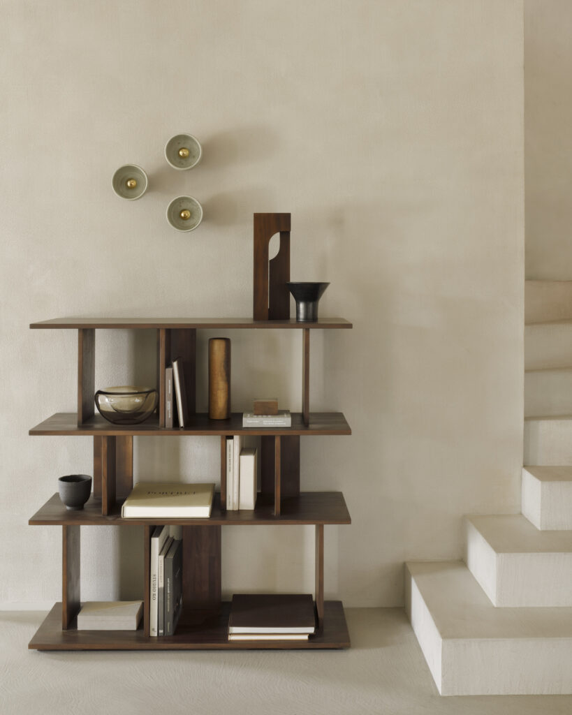 Une bibliothèque en teck vernis avec divers objets et livres se dresse contre un mur beige. Trois appliques murales décoratives sont montées au-dessus et un escalier blanc est visible à droite. La finition marron ajoute de la chaleur et du caractère à l'espace.