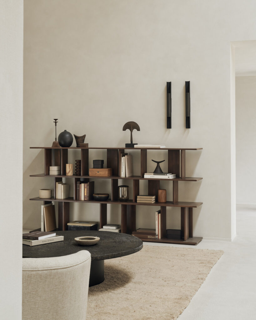 Un salon moderne comprend une étagère irrégulière en teck verni, remplie de livres et d'objets de décoration, une table basse ronde et un tapis beige. Les murs de couleur claire sont rehaussés de deux appliques murales noires.