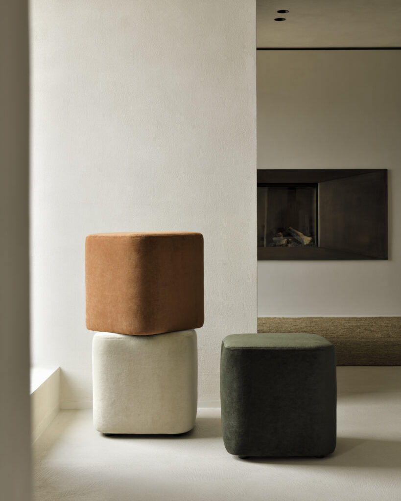 Trois poufs cubes sont empilés dans un salon moderne et minimaliste. Les poufs, fabriqués à partir de tissu cannelle, sont de couleur marron, blanc et vert foncé. L'arrière-plan présente un mur avec une cheminée encastrée.
