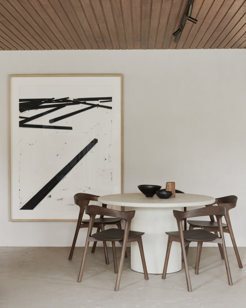 Une table à manger ronde blanc cassé avec quatre chaises en bois est placée contre un mur avec une grande peinture abstraite en noir et blanc. Le plafond présente d'élégants panneaux de bois.