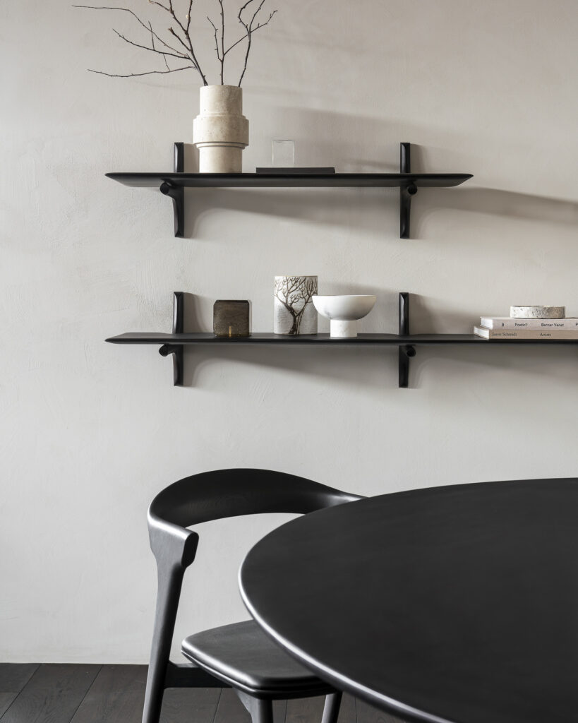 Coin repas minimaliste avec table ronde noire, chaise noire et deux étagères murales marron foncé ornées d'objets décoratifs, de livres et d'un vase avec des branches sur un mur de couleur claire.