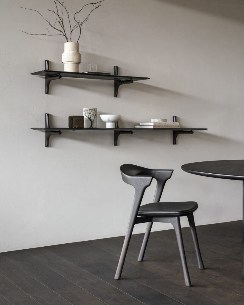 Chambre minimaliste avec une chaise et une table en bois sombre, complétées par deux étagères murales PI contenant des objets de décoration simples, notamment un vase, une tasse et des petits livres, le tout contre un mur gris clair et un parquet en bois foncé. La finition en acajou verni du meuble ajoute de l'élégance à l'espace.