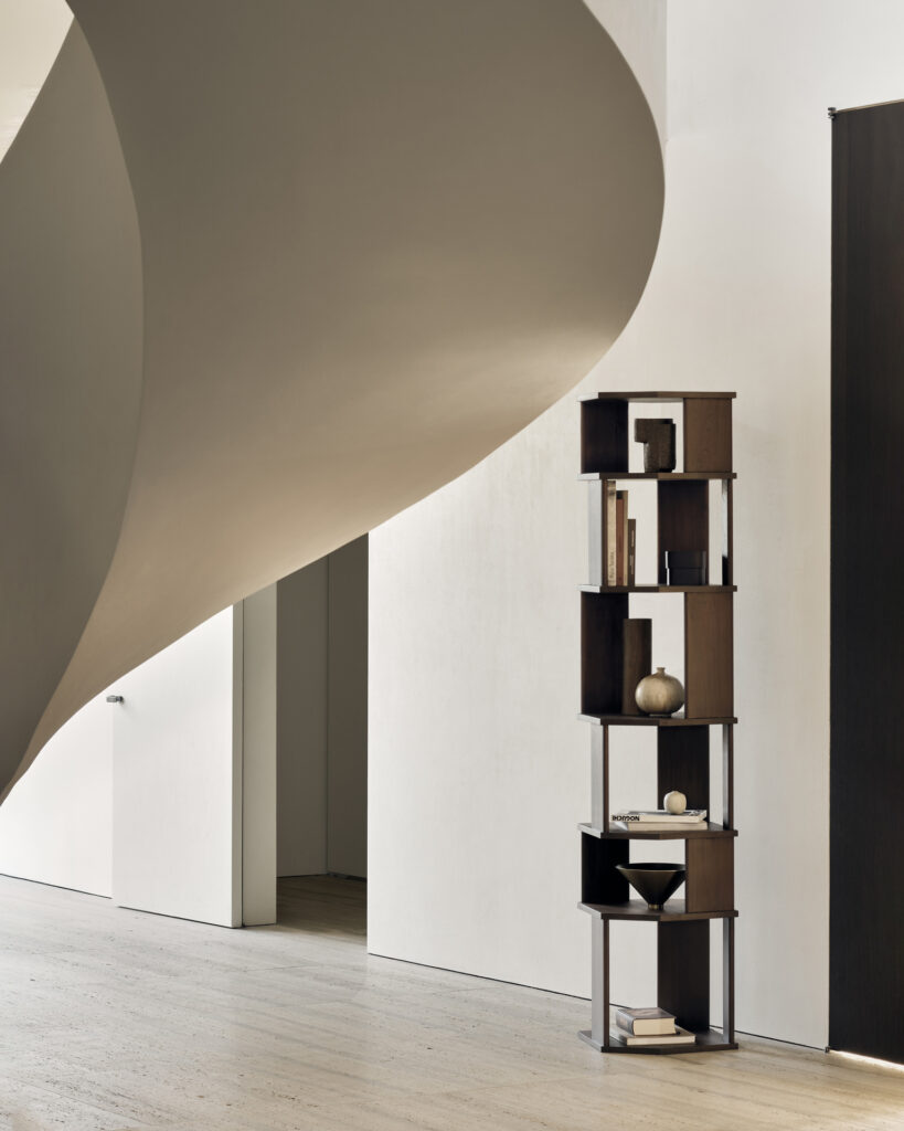 Une bibliothèque haute et étroite en bois teintée d'un riche vernis brun, remplie de divers objets de décoration et de livres, se dresse contre un mur blanc à côté d'un élément architectural aux lignes douces et courbes.