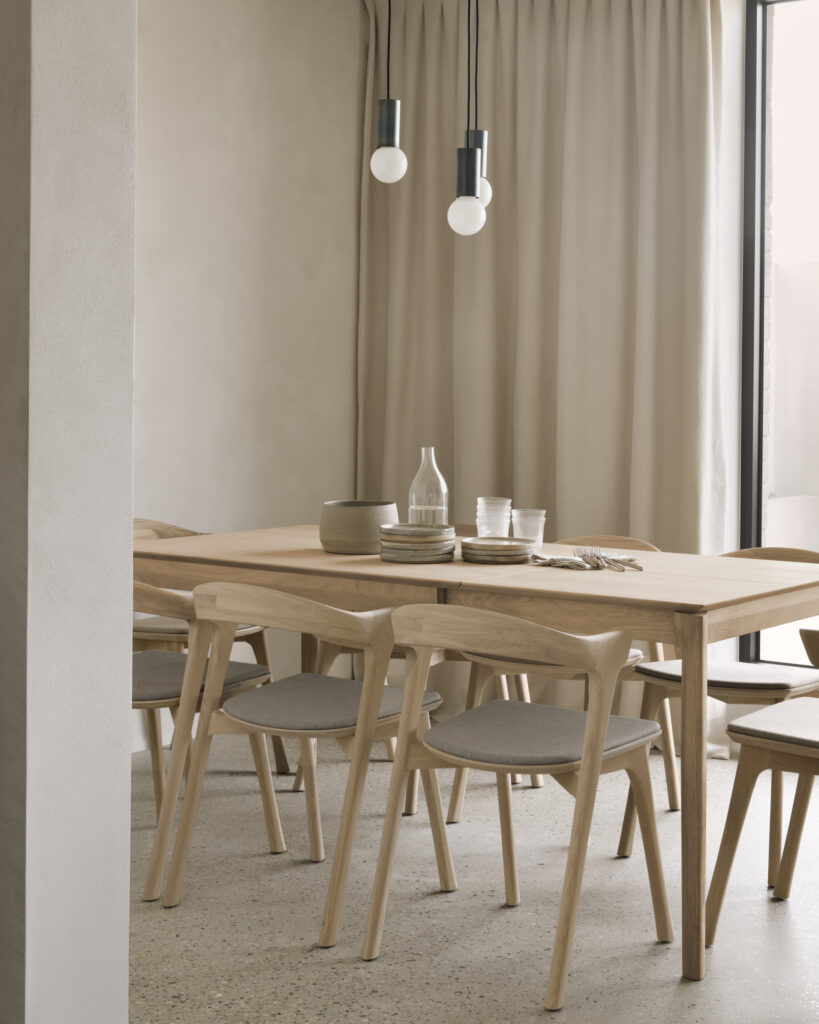 Une salle à manger minimaliste avec une table Bok en bois, six chaises, une paire de suspensions et des rideaux de couleurs neutres. La table est dressée avec une carafe, des tasses et une pile d'assiettes. Cette pièce polyvalente est dotée d'une rallonge en chêne l'étendant de 120 à 180 cm, parfaite pour divers rassemblements.