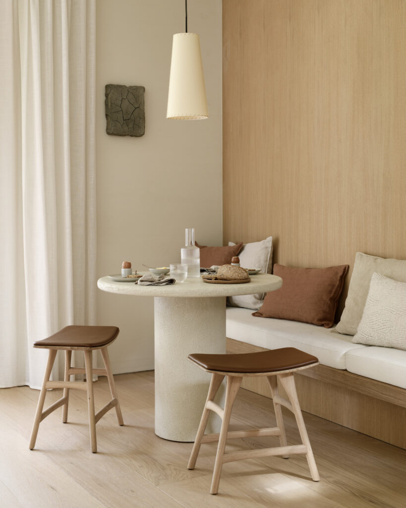 Un coin repas minimaliste avec la table ronde en pierre Table Elements, deux tabourets en bois à l'assise brune, un banc encastré avec des coussins neutres et une lampe suspendue au dessus de la tête.