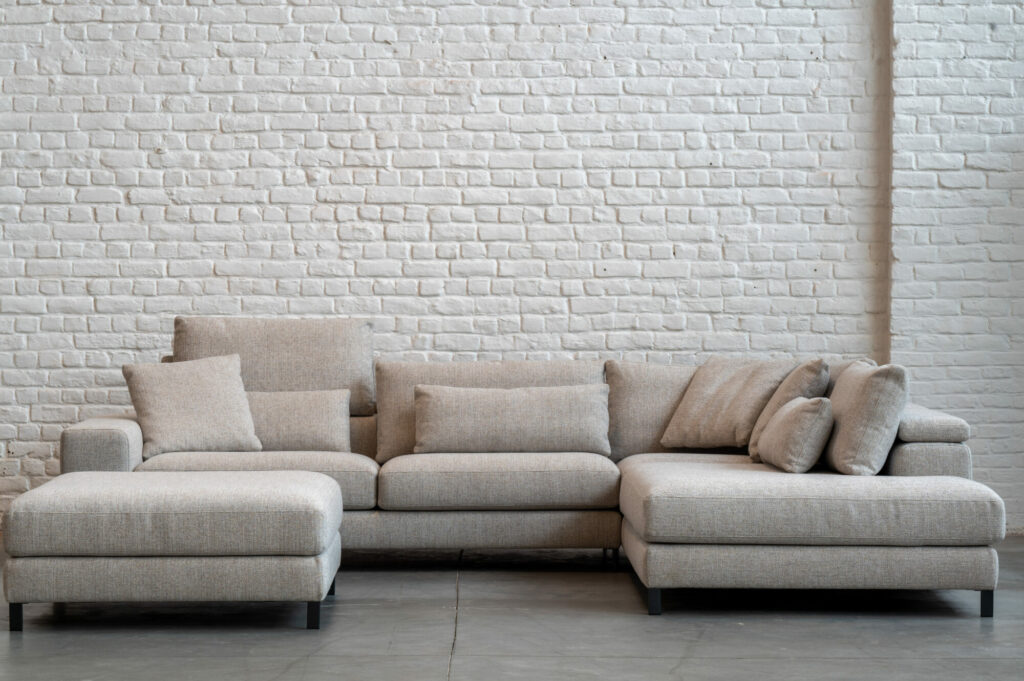 Le canapé sectionnel Horizon [3,5z +L2Hz] - C50 Reserve 4192 avec plusieurs coussins est élégamment positionné contre un mur de briques blanches sur un sol en béton, offrant un havre de confort douillet.
