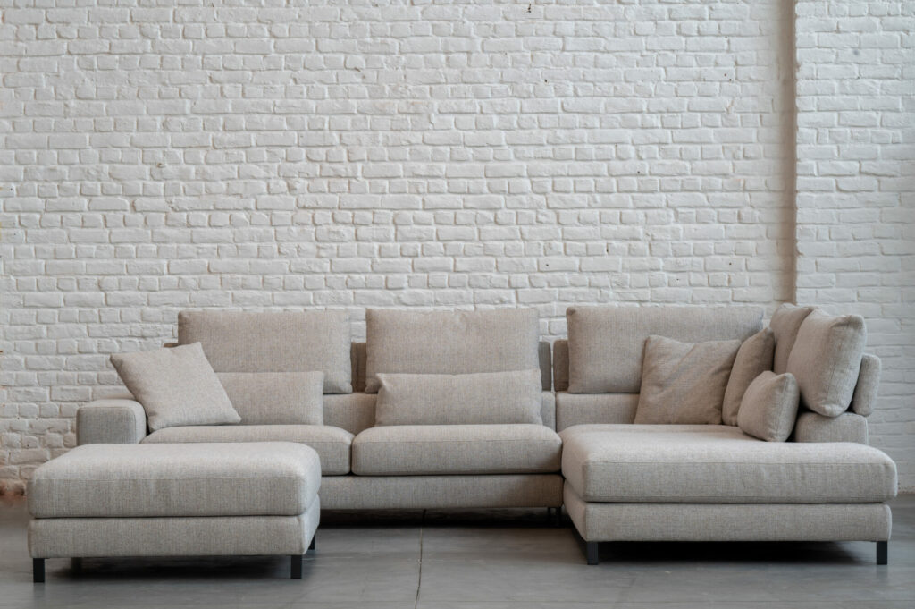 Un canapé sectionnel beige, inspiré de l'élégance de l'Horizon [3,5z +L2Hz] - C50 Reserve 4192 - pieds col, avec plusieurs coussins est placé contre un mur de briques blanches sur un sol en béton.