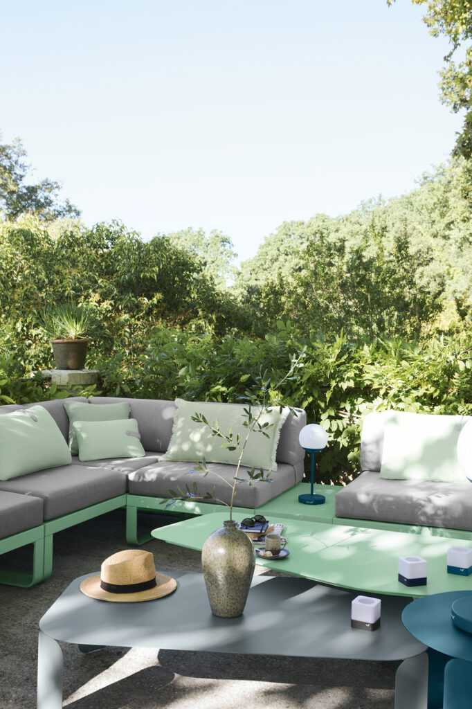 Terrasse extérieure moderne avec un canapé sectionnel gris et vert menthe, la Table basse Bebop rectangulaire, un vase décoratif, des petites lampes et un chapeau de paille, le tout entouré d'un feuillage vert luxuriant.