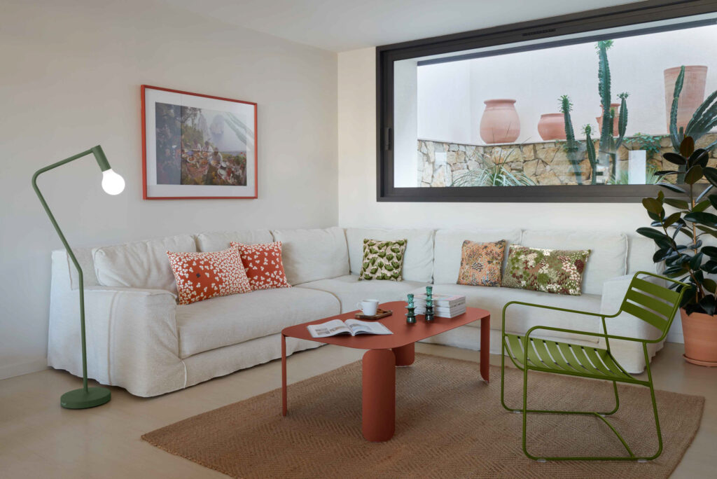 Salon contemporain comprenant un canapé sectionnel blanc, un lampadaire vert, une table basse rectangulaire Bebop, des coussins à motifs et une grande fenêtre avec vue sur les plantes extérieures et les pots en terre cuite.