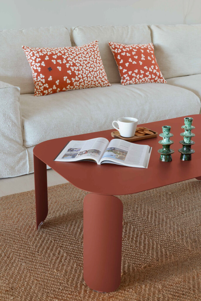Une table basse Bebop rectangulaire de couleur rouille accueille un magazine ouvert, une tasse blanche sur une soucoupe et des bougeoirs verts, placés devant un canapé beige avec des coussins à motifs.
