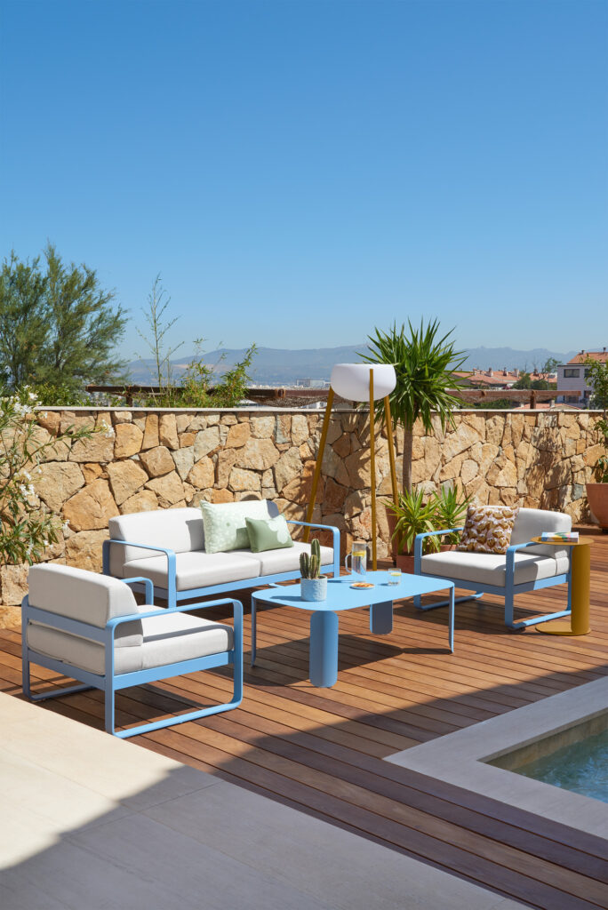Terrasse extérieure moderne avec mobilier bleu et blanc, une table basse Bebop rectangulaire contenant des boissons et des plantes, terrasse en bois, mur en pierre et un ciel clair en arrière-plan.