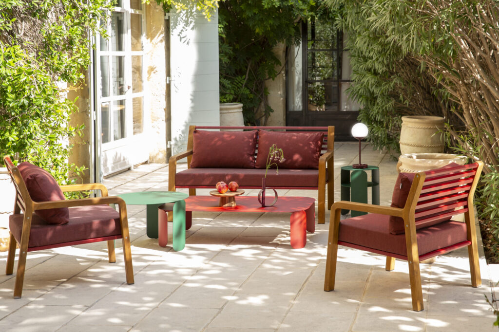 Ensemble de patio extérieur avec chaises en bois, coussins marron, une table basse Bebop rectangulaire rouge, une table verte et une petite lampe, entouré de plantes et d'un sol en pierre à côté d'un bâtiment.