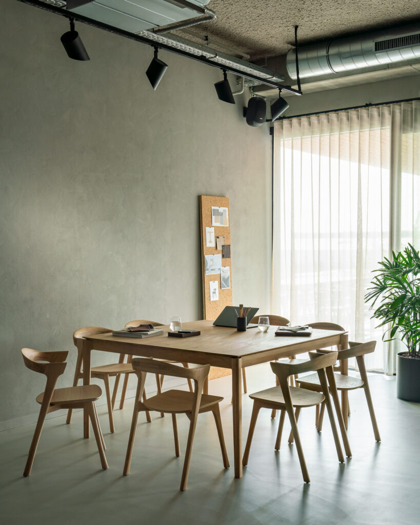 Une salle de réunion moderne avec une table en chêne, des chaises en bois et un verre d'eau sur la Table Bok. Un tableau en liège avec des papiers orne le mur et une grande plante en pot se dresse près de la fenêtre avec des rideaux transparents.