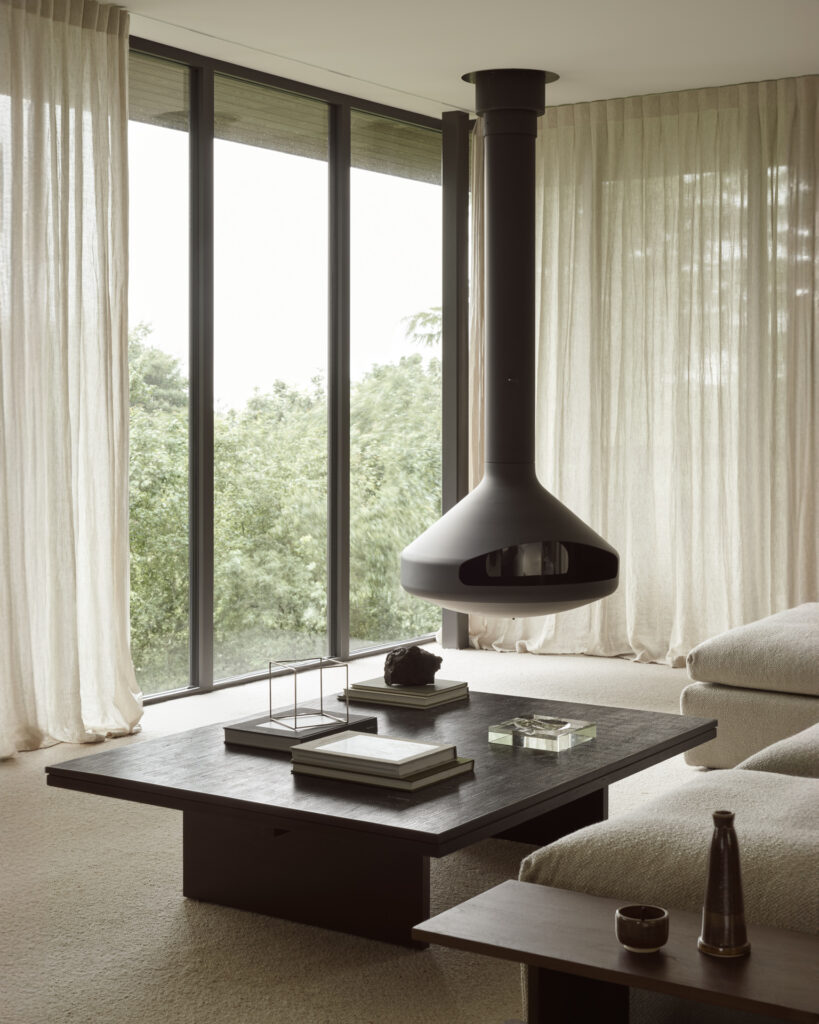 Un salon moderne comprend une cheminée suspendue, de grandes fenêtres en verre avec des rideaux lumineux, la table basse Grooves avec de subtiles textures de bois foncé et un canapé sectionnel beige.