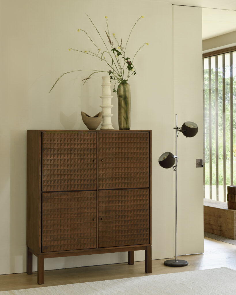 Une armoire Sono en bois ornée de vases et d'un bol se dresse contre un mur clair, rehaussé d'un lampadaire moderne dans une pièce lumineuse aux grandes fenêtres.