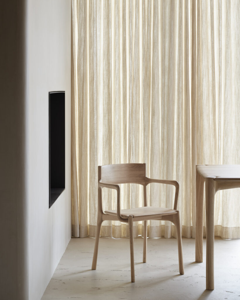 Une pièce minimaliste comprend une chaise de salle à manger PI avec accoudoirs et une table en bois assortie à côté de rideaux beiges transparents. Un rectangle noir creux est encastré dans le mur blanc à gauche, et le sol est de couleur claire.