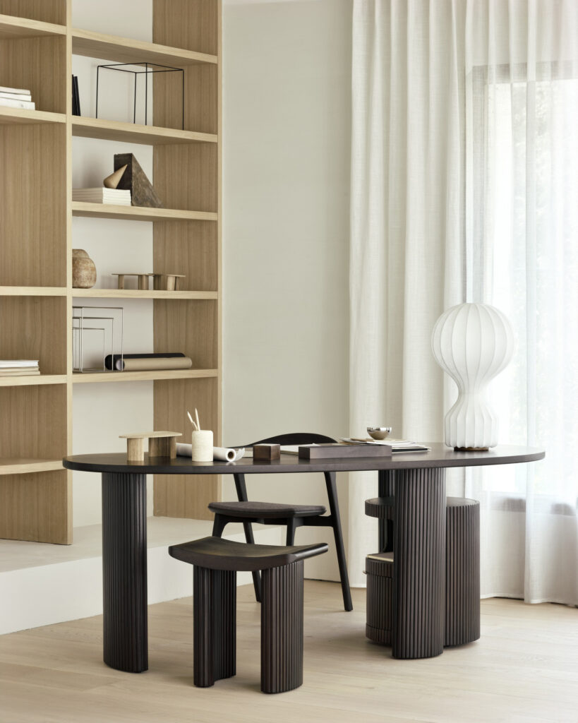 Un bureau moderne est composé d'un bureau en bois foncé avec des chaises assorties, d'une décoration minimaliste, d'œuvres d'art, d'une lampe blanche texturée et de l'élégant meuble à tiroirs Boomerang. La lumière filtre à travers des rideaux blancs transparents à côté des étagères intégrées.