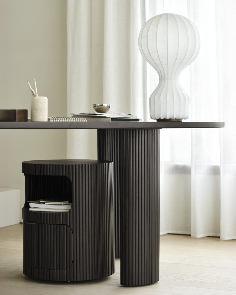 Un bureau moderne comprenant une table cylindrique sombre, un bloc-tiroirs Boomerang, un cahier, un pot à stylos et une lampe blanche en forme de boomerang près d'un rideau blanc.