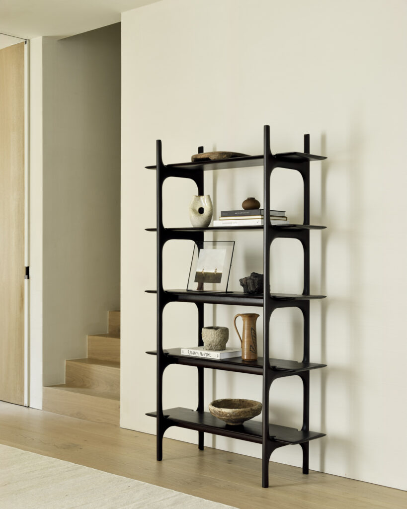 Un support PI noir contre un mur beige présente des objets décoratifs, des livres et un petit vase dans une pièce minimaliste et moderne avec une ambiance de rangement industrielle, avec du parquet et des escaliers en arrière-plan.