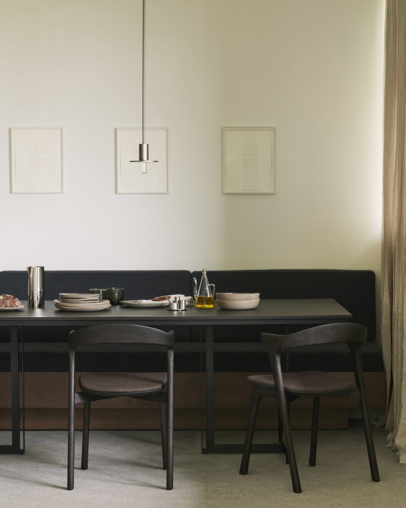 Une salle à manger minimaliste est mise en valeur par une table à manger Tacet sombre, associée à des chaises noires, une suspension moderne et des œuvres d'art murales neutres. La table est dressée avec deux couverts et ornée de quelques ustensiles de cuisine. Une fenêtre de la pièce est drapée de rideaux de couleur crème.