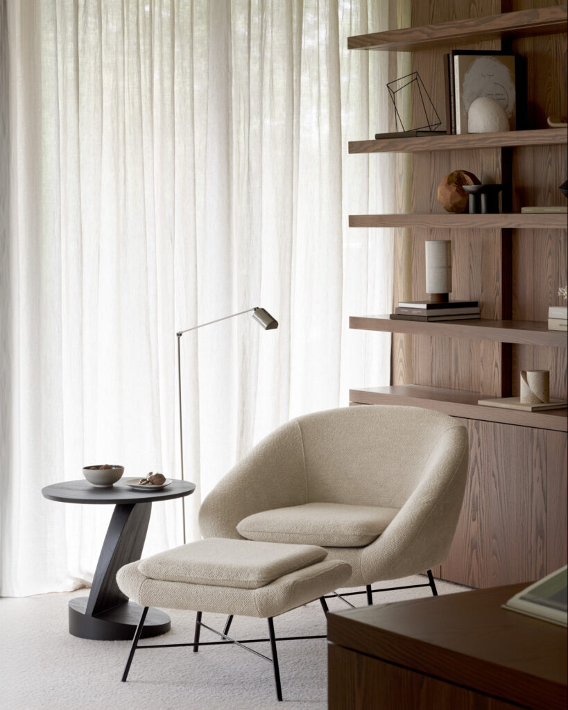 Un coin lecture moderne avec une chaise beige associée au repose-pieds Barrow, une table d'appoint noire et un lampadaire fin. Des objets décoratifs et des livres ornent les étagères en arrière-plan, complétées par de longs rideaux transparents.