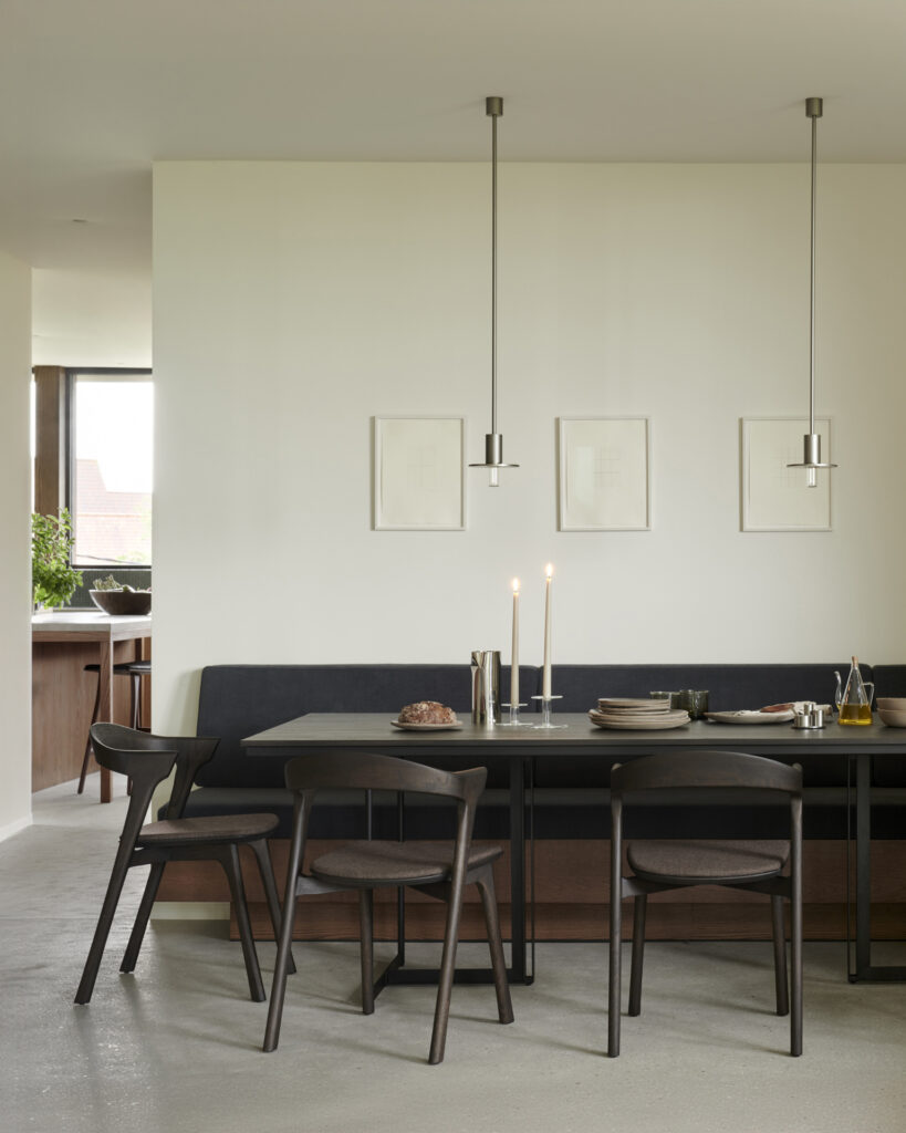 Un espace salle à manger élégant avec une table rectangulaire sombre et épurée complétée par quatre chaises de salle à manger Bok et un banc assorti. L'aménagement comprend la lueur chaleureuse de deux bougies allumées et des décorations minimalistes, le tout sur fond de murs de couleur claire ornés d'œuvres d'art et éclairés par un éclairage suspendu.