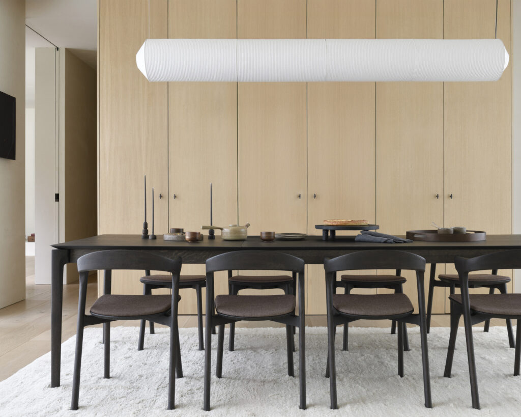 Une salle à manger minimaliste avec une table à manger extensible Bok en Chêne teinté brun avec une finition vernis, six chaises marron assorties et un grand luminaire cylindrique au-dessus. Le mur derrière est doté de panneaux et d'armoires en bois, avec un tapis blanc en dessous.