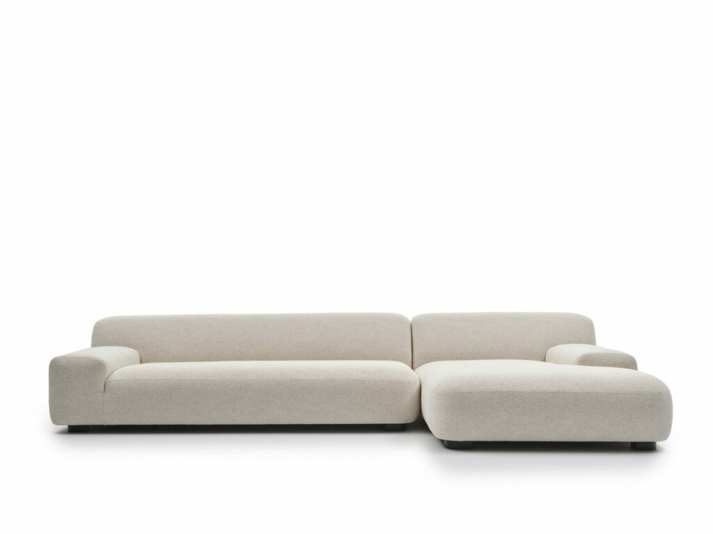 Canapé moderne en L de couleur beige avec dossier bas et accoudoirs, avec une chaise longue sur le côté droit. Le canapé est recouvert de tissu doux et se détache sur un fond blanc uni.
