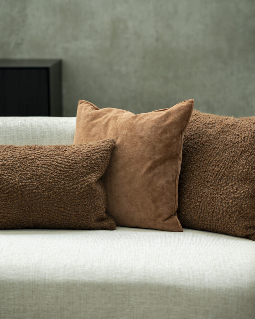 Un canapé beige avec trois coussins marron, dont un en daim et un autre en forme de vague avec un tissu texturé, est posé contre un mur de couleur neutre. Le coussin en forme de vague présente un motif subtil, ajoutant une touche élégante à l'atmosphère sereine.