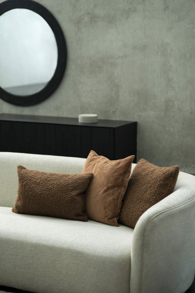 Un canapé beige clair avec deux coussins marron et un coussin Wave se trouve devant une console en bois foncé. Un miroir rond est suspendu au-dessus de la console contre un mur gris texturé.